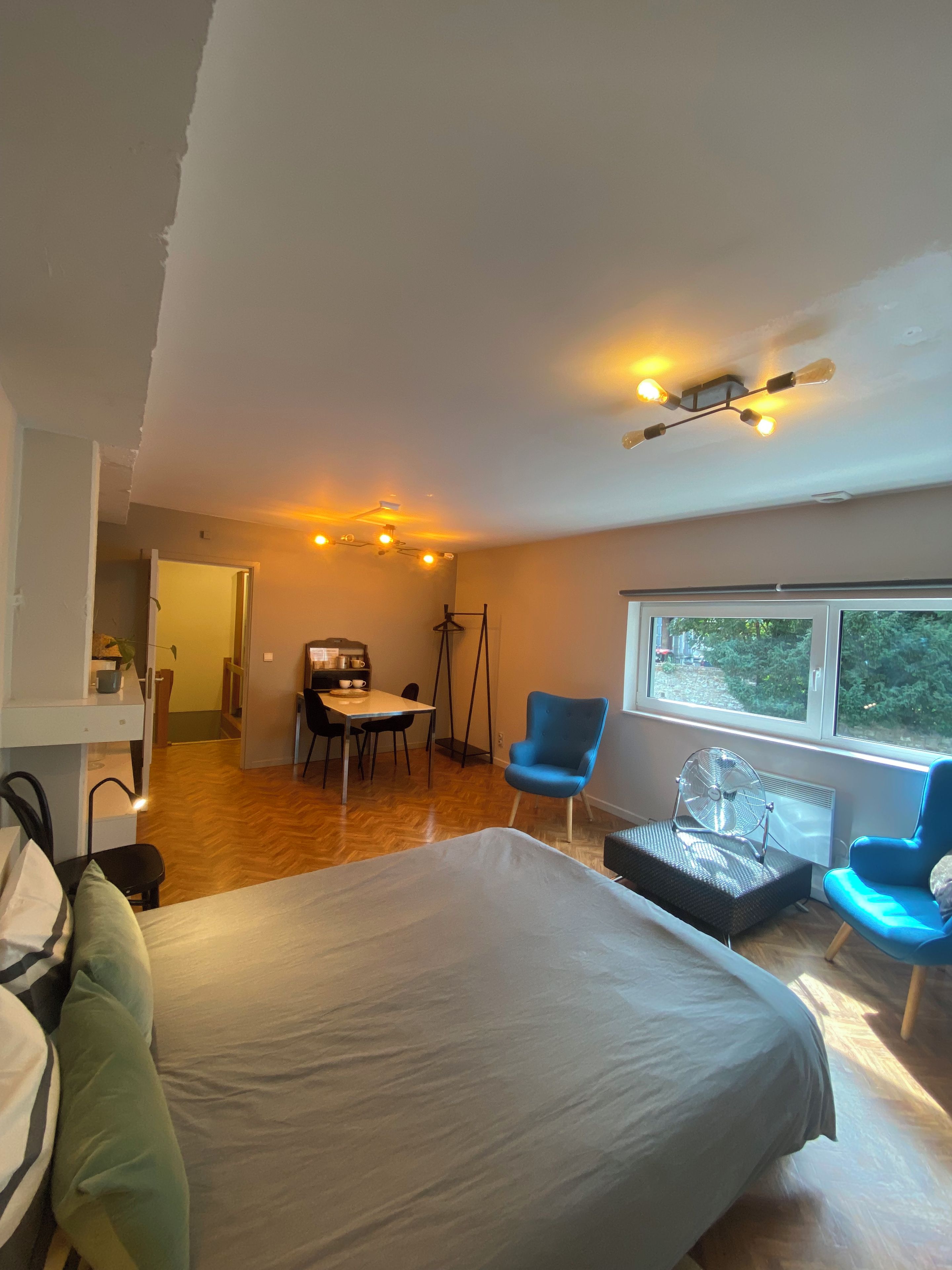 A bedroom with a bed, a desk, two chairs, a fan, and a window with a view.
