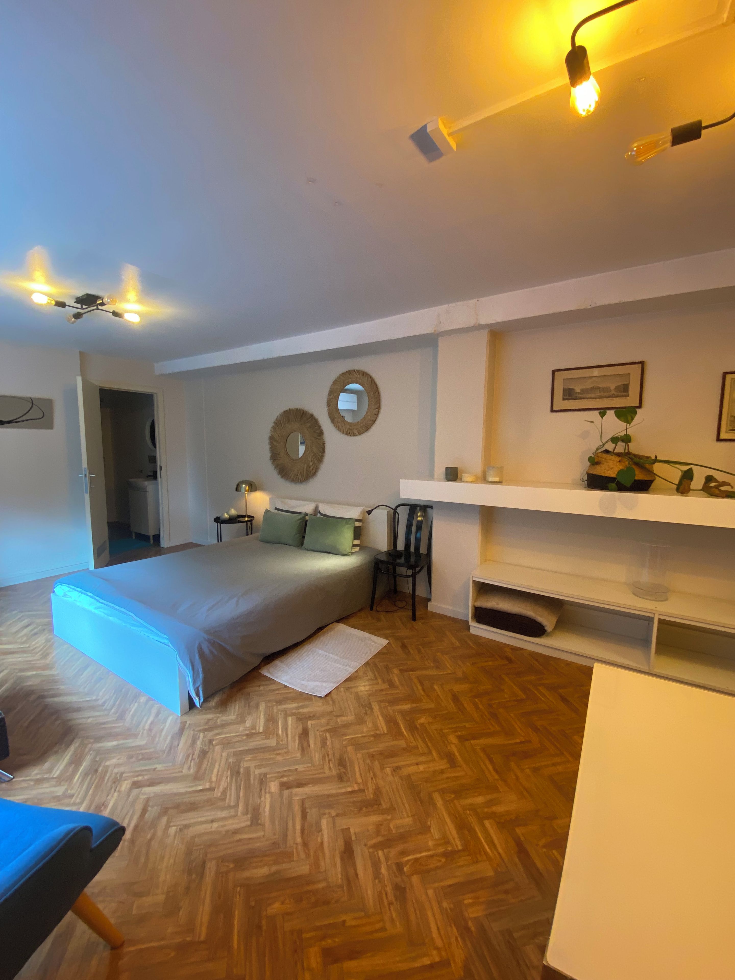 Ein Schlafzimmer mit einem Bett, Nachttischen, einem Sessel, einem Regal mit Dekorationen und einem Fischgrätparkett.