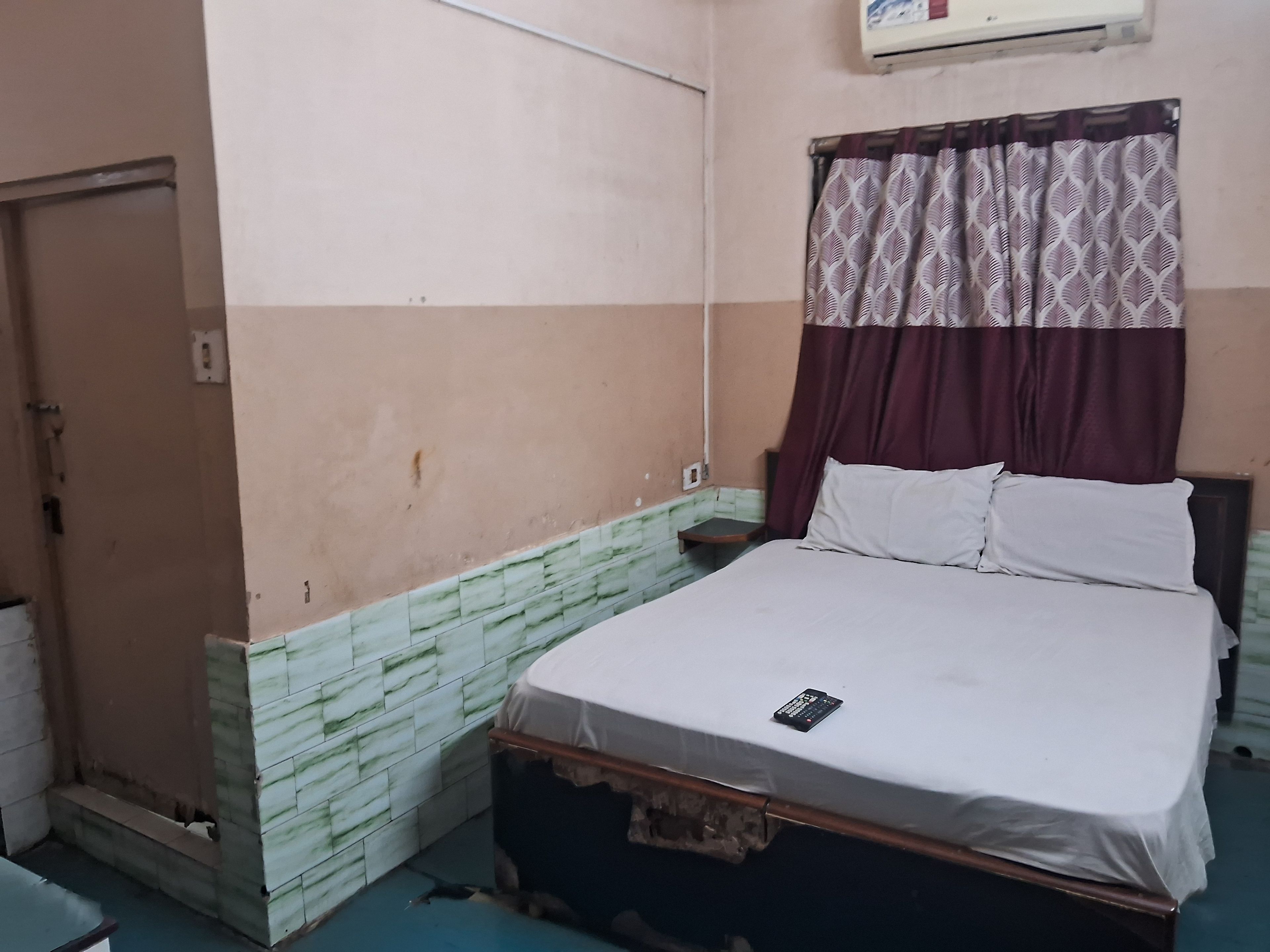 Un dormitorio con cama, aire acondicionado de pared, cortina estampada y parte baja de pared con azulejos.