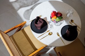 Une table de salle à manger ronde, recouverte d’une nappe blanche à motifs, avec des assiettes noires, des couverts dorés et un vase rempli de fleurs.