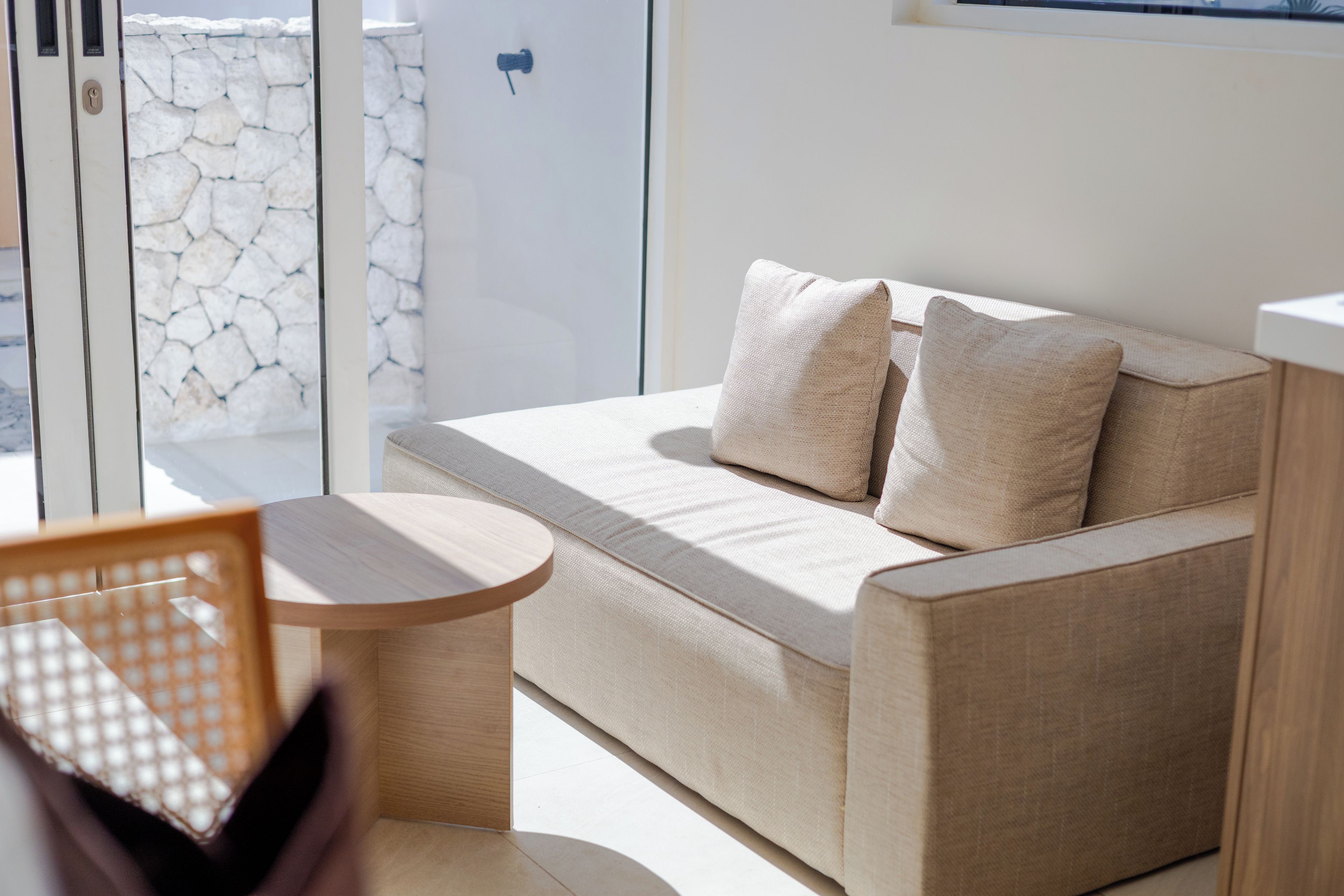 Een moderne woonkamer met een beige bank, twee kussens, een kleine houten tafel en uitzicht op een stenen muur door een glazen deur.