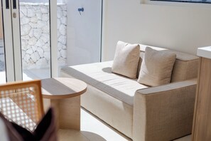Un salon moderne avec un canapé beige, deux coussins, une petite table en bois et une vue sur un mur de pierre, visible à travers une porte vitrée.