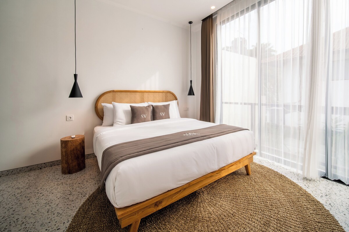 Una habitación de hotel moderna con una cama grande, dos lámparas de noche, una mesita de noche de madera y vistas a la ciudad a través de cortinas translúcidas.
