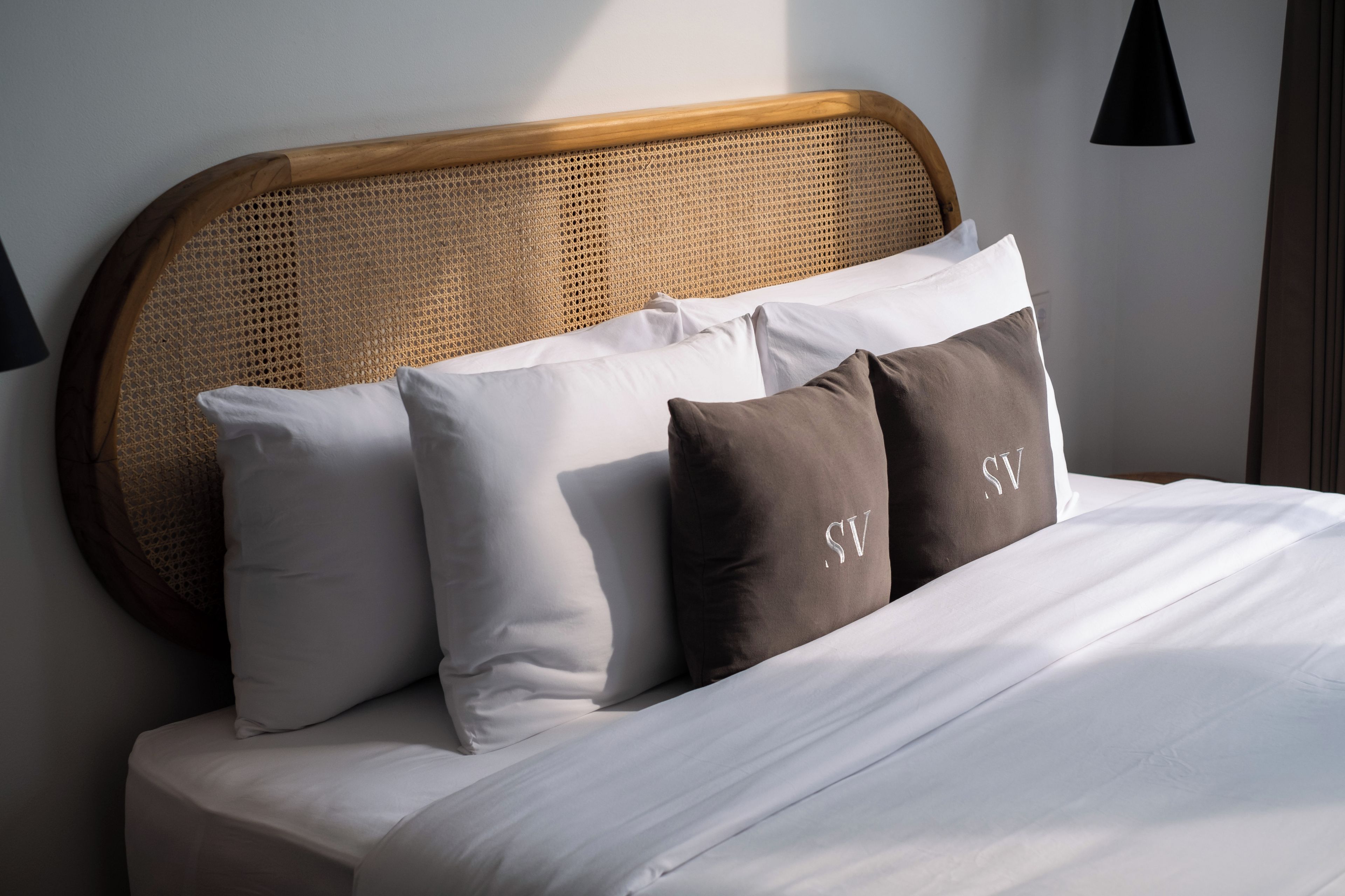 Una cama con ropa blanca, cabecera tejida y dos almohadas con un monograma.
