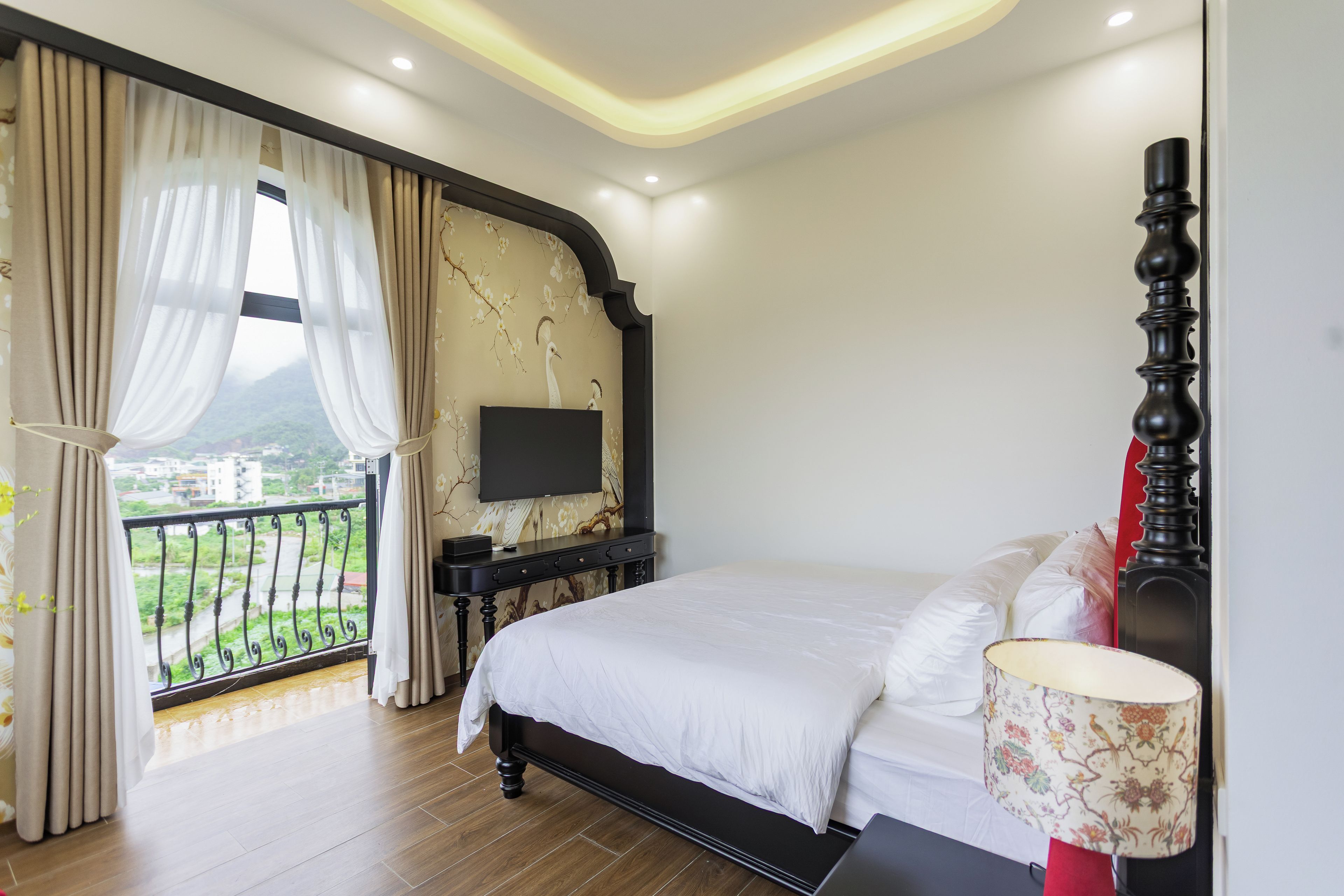 A bedroom with a bed, a TV on a stand, a balcony with curtains, and a view of greenery.