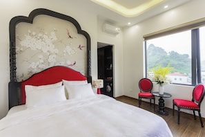 A hotel room with a large bed, a red upholstered chair, a small table with a vase, and a window with a view of greenery and buildings.