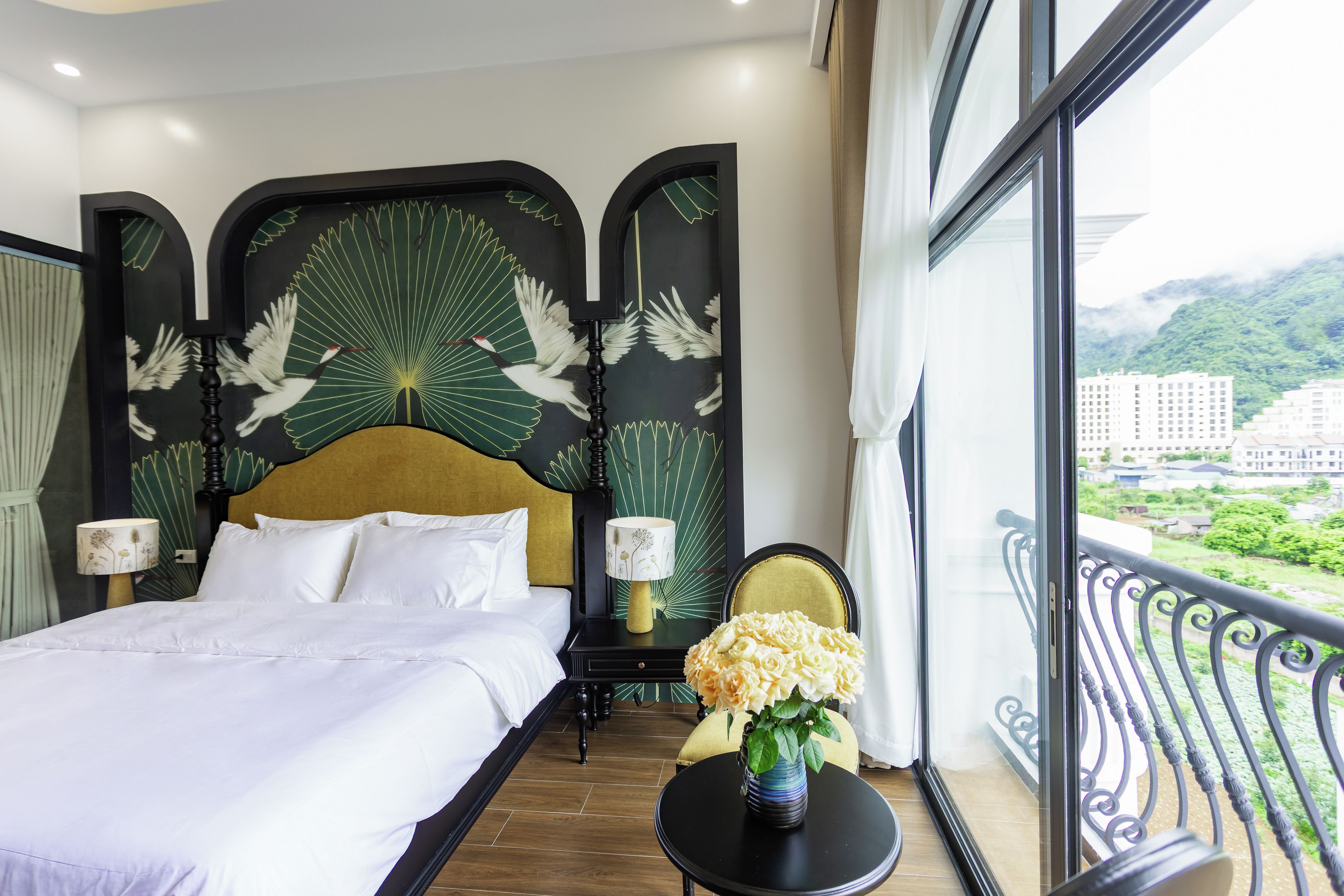 A hotel room with a large bed, two bedside tables, a chair, a small table with flowers, and a balcony with a view of buildings and greenery.