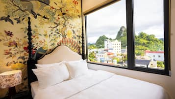 Una cama con sábanas blancas, cabecera y ventana con vistas a un paisaje urbano y zonas verdes.