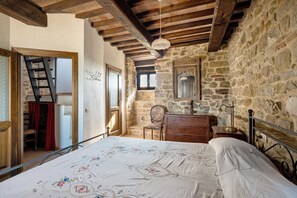 A bedroom with a stone wall, wooden beams, a bed with a floral bedspread, a dresser, a chair, and a ladder leading to a door.