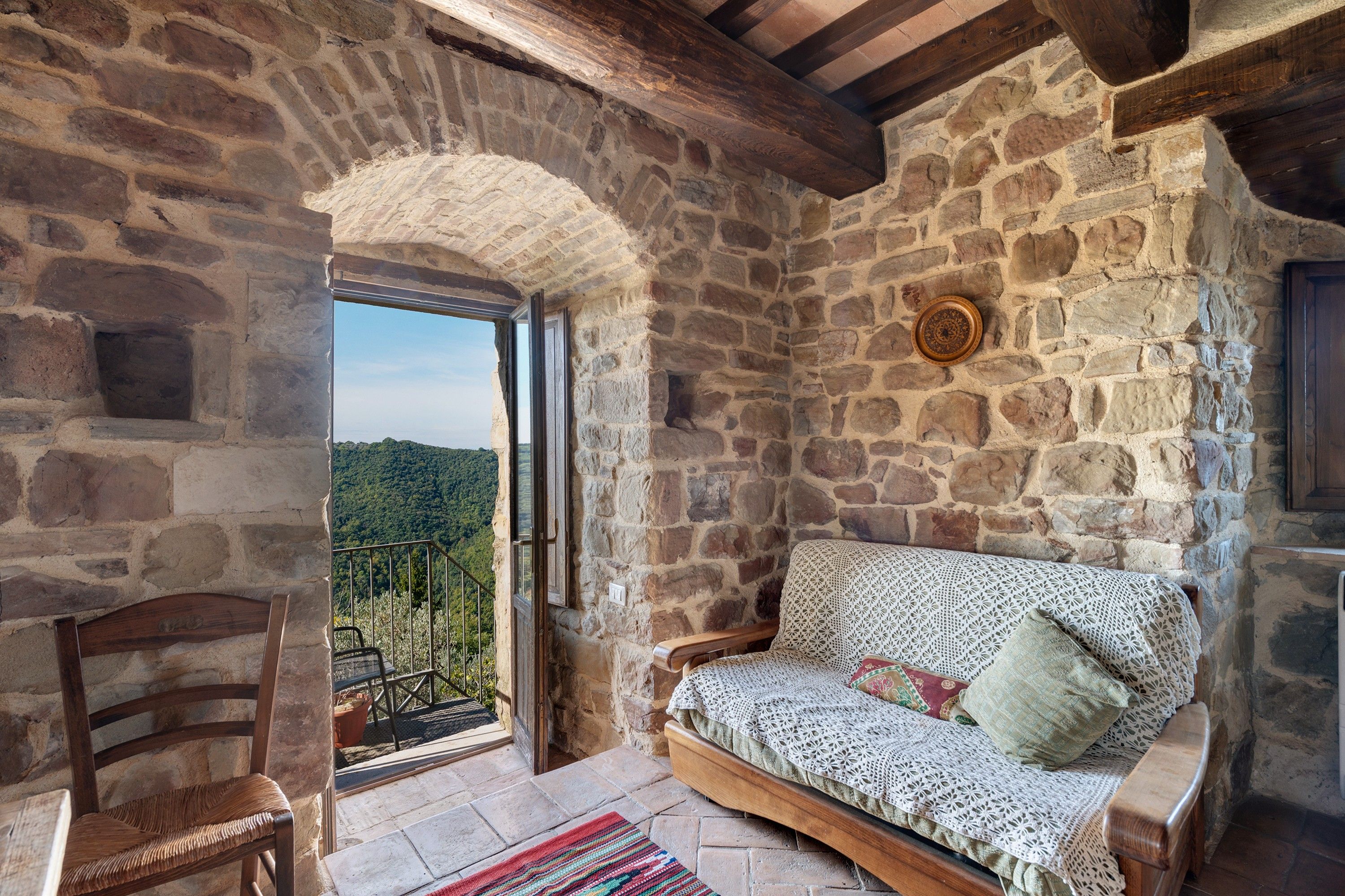 Ein Zimmer mit Steinwänden, einer Holzbank, einem gemusterten Sofa und Blick auf eine bewaldete Landschaft.