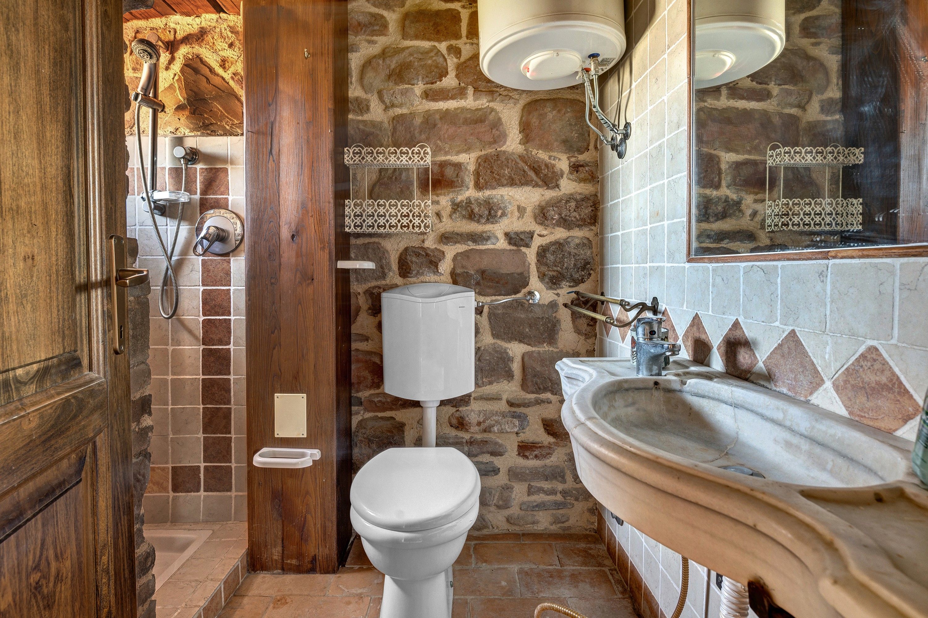 Une salle de bain avec un mur en pierre, une porte en bois, des toilettes blanches et un lavabo surmonté d’un miroir.