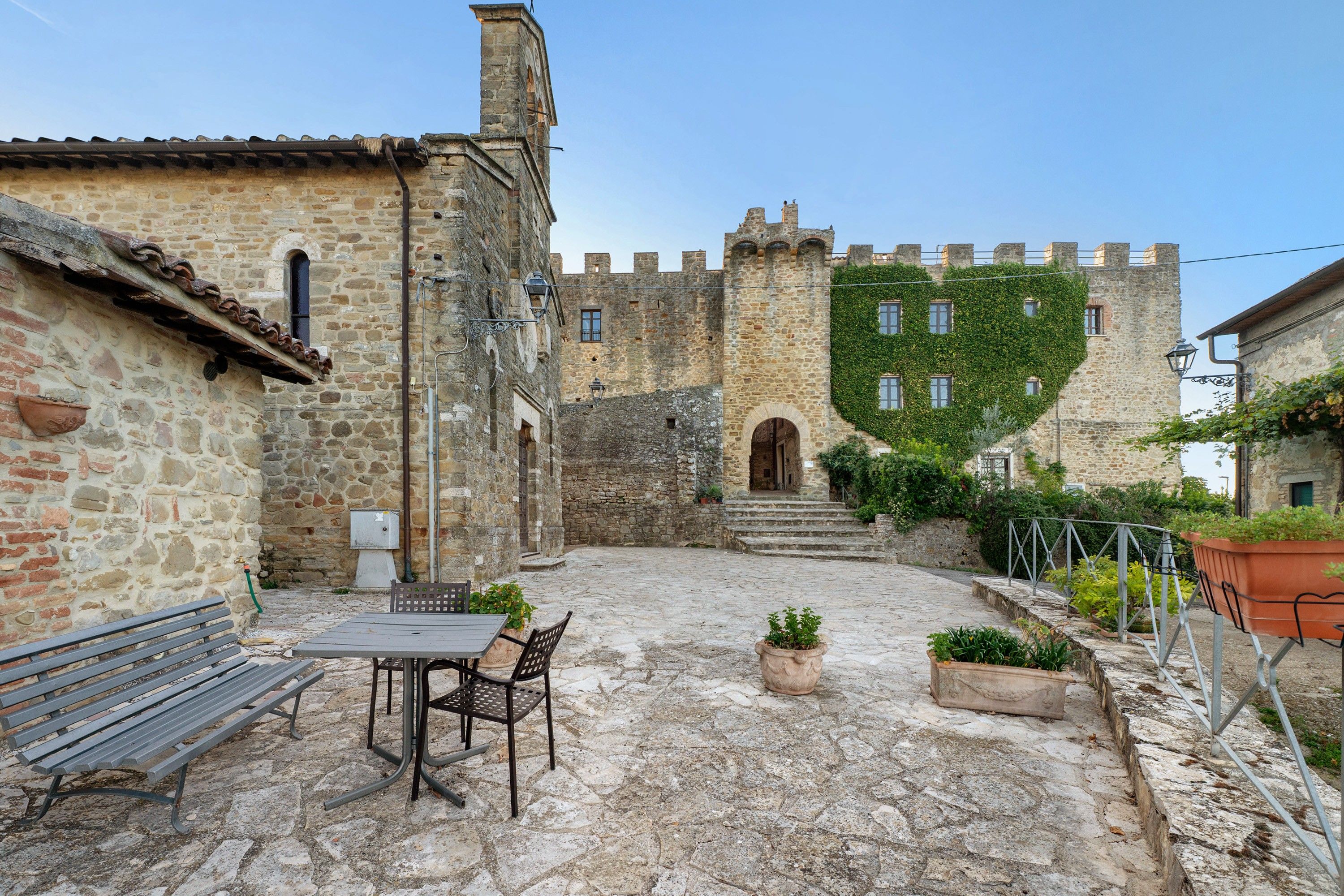 A courtyard with stone walls, a bench, a table, and potted plants.