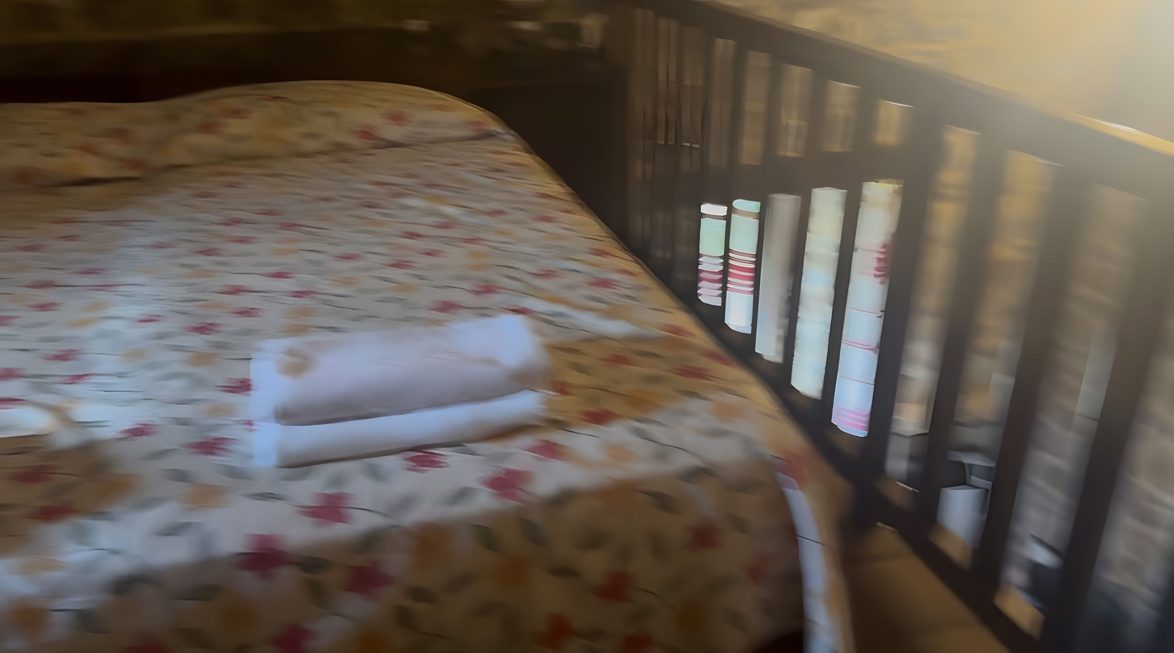 A bed with a floral bedspread and a rolled towel, a wooden headboard, and a window with vertical blinds.