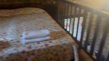 A bed with a floral bedspread and a rolled towel, a wooden headboard, and a window with vertical blinds.