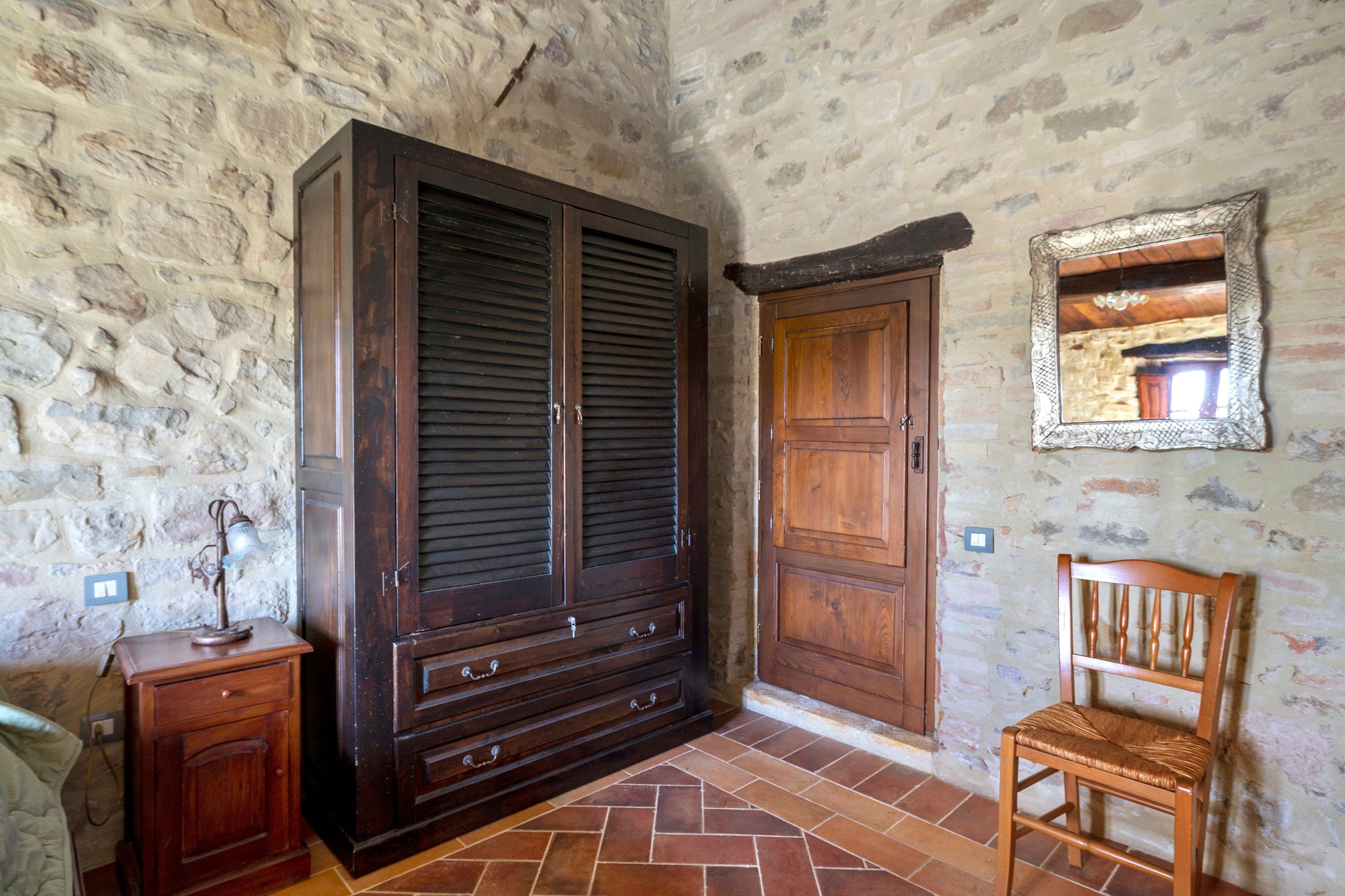 Una habitación con un armario grande de madera, una silla de madera, una mesita con una lámpara y un espejo en la pared.