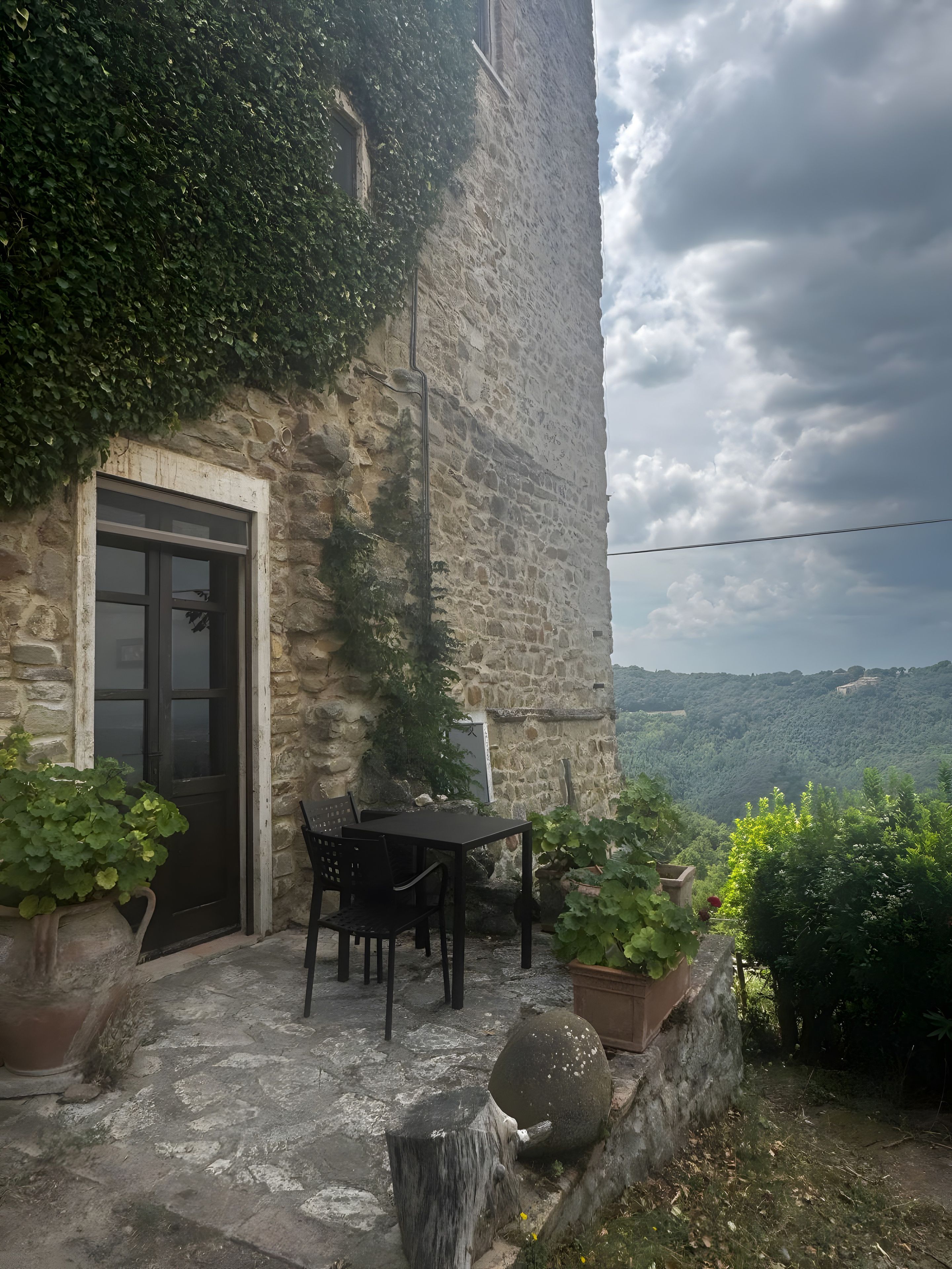 A stone building with a door and window, outdoor seating with chairs and a table, and a view of a hilly landscape.