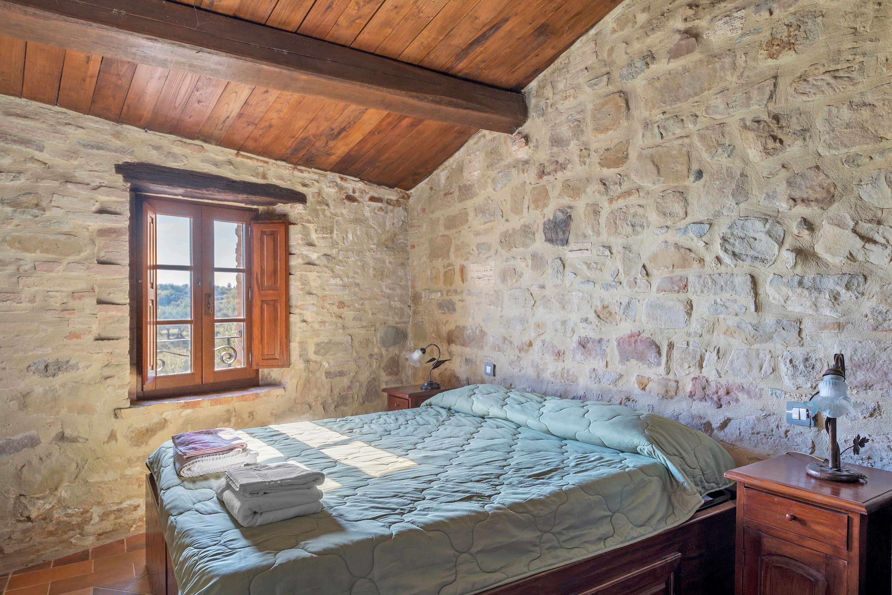 Ein Schlafzimmer mit einer Steinwand, einer Holzbalkendecke, einem Bett mit Quilt, einem Nachttisch mit Lampe und einem Fenster mit Holzläden.