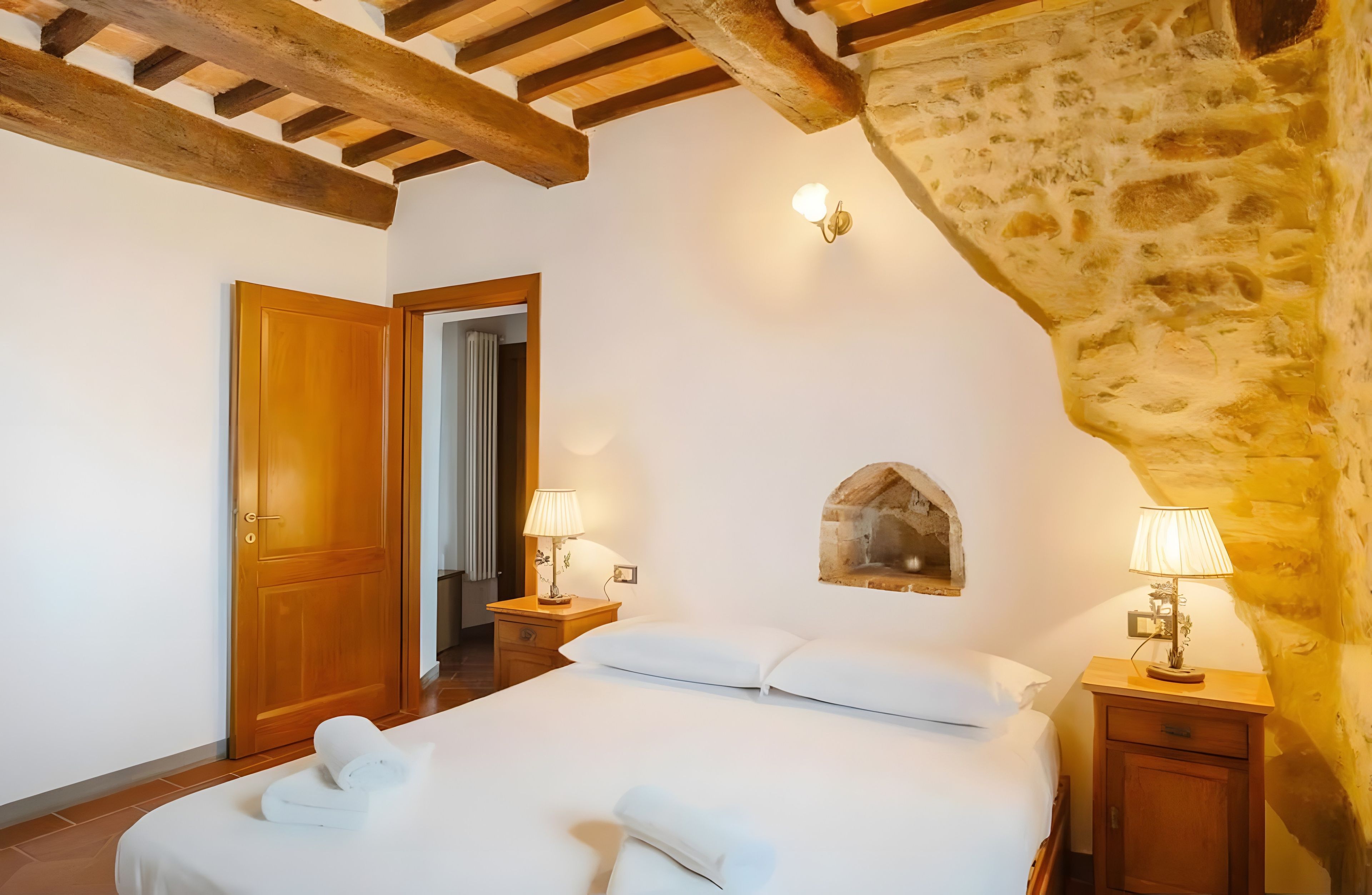 Una camera da letto con soffitto in legno, parete in pietra, un letto con lenzuola bianche, due abat-jour e un piccolo quadro.