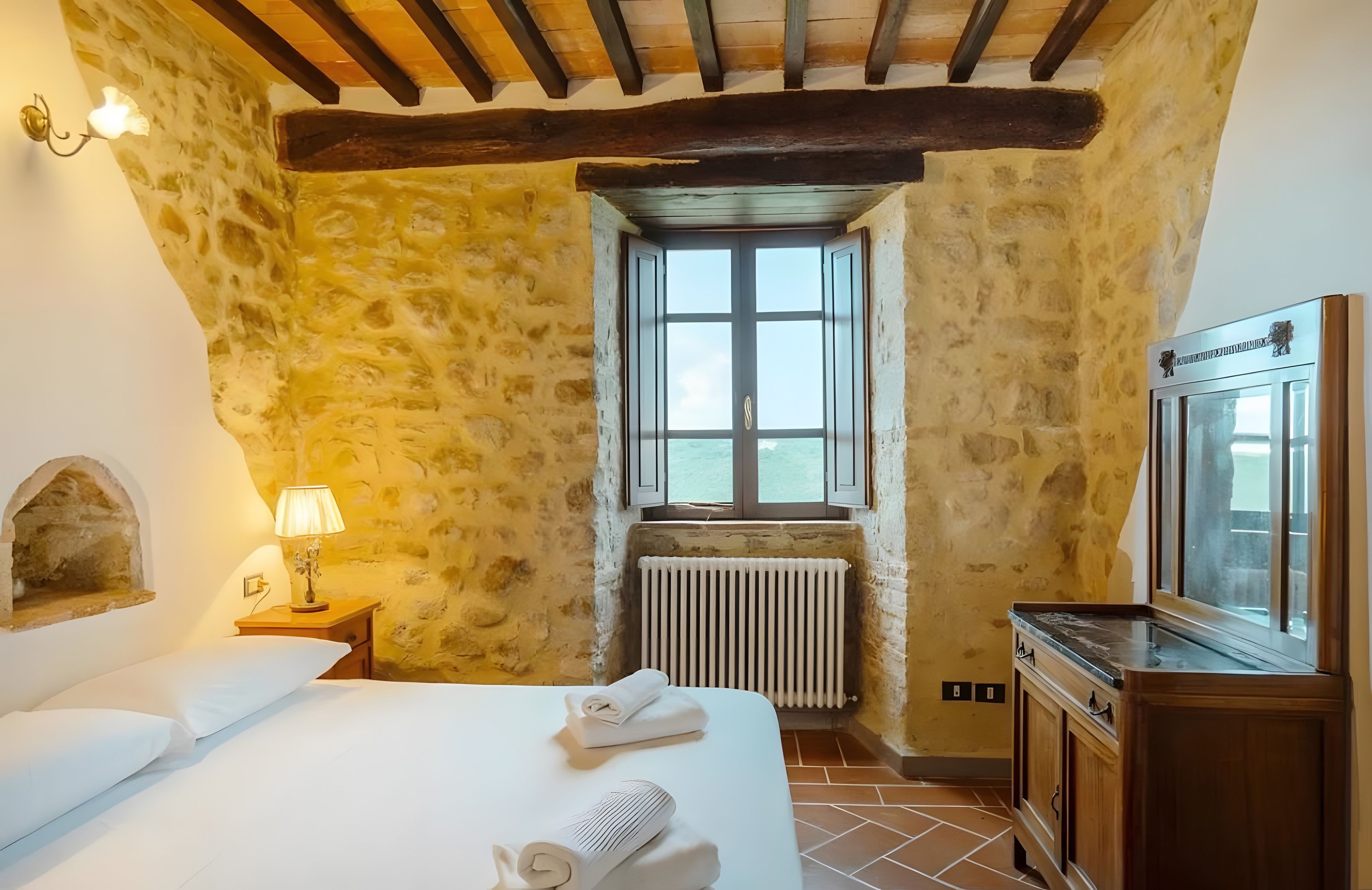 Una camera da letto con parete in pietra, un letto con lenzuola bianche, un termosifone, una finestra vista e un armadio in legno.