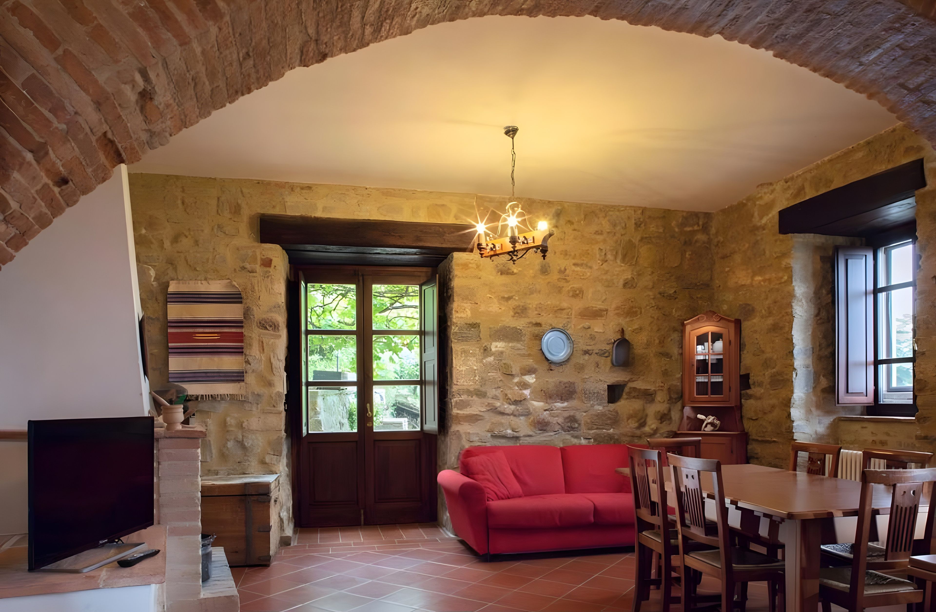 A rustic interior with a stone wall, a red sofa, a wooden dining table, and a chandelier.
