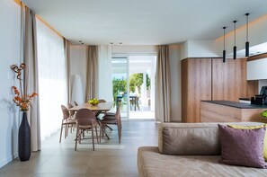 A modern living room with a sofa, dining table, and chairs. There is a kitchen area with wooden cabinets and a countertop. A large window provides a view of the outdoors. - Luxurious villa with outdoor pool, heated indoor pool, wellness area, and outdoor Jacuzzi in Cornino – DiCasaInSicilia (Custonaci)