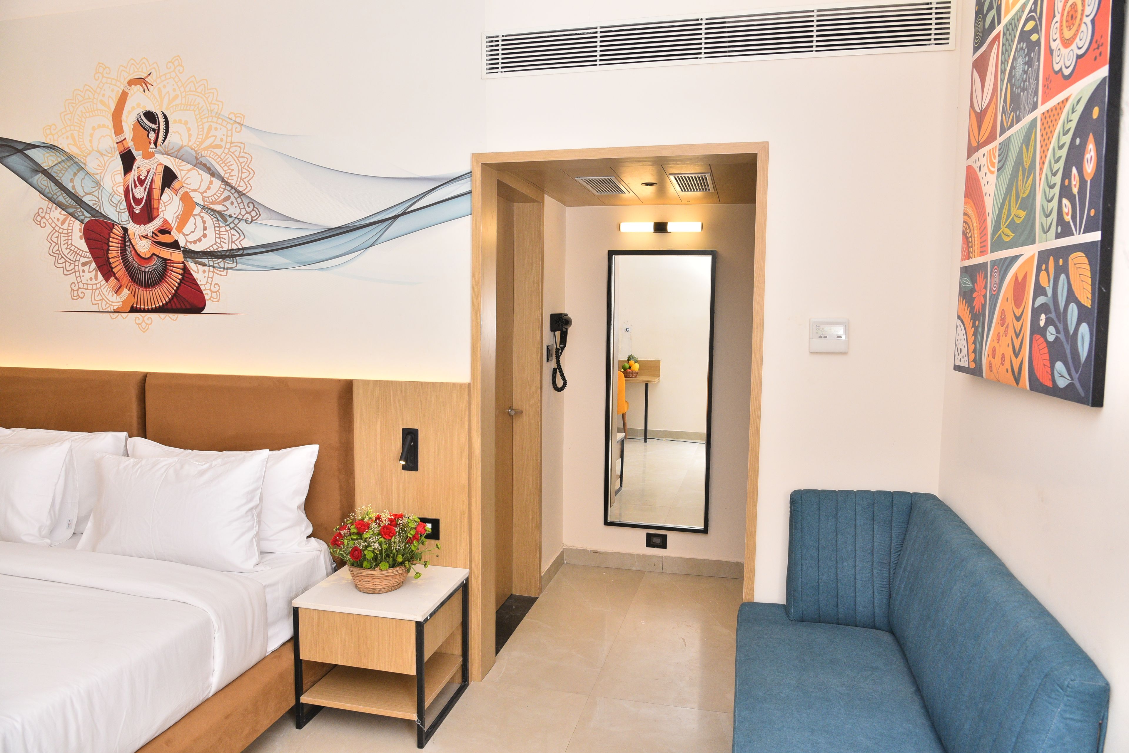 Een hotelkamer met een bed, een klein tafeltje met een bloemstuk, een blauwe bank, een spiegel en een muur met een kleurrijk abstract ontwerp.