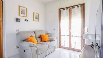 Ein Wohnzimmer mit einem beigen Sofa, zwei orangefarbenen Kissen, einer Holzkommode und einer Schiebetür zum Außenbereich.