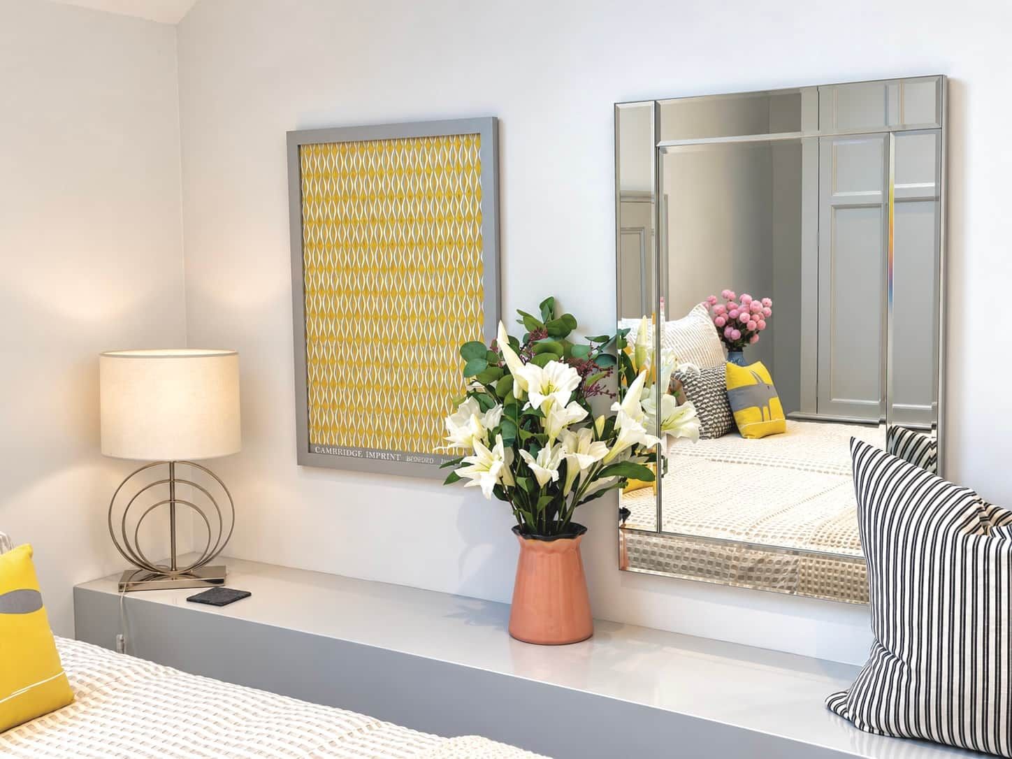 A modern bedroom with a bed, bedside table, lamp, mirror, and decorative items.