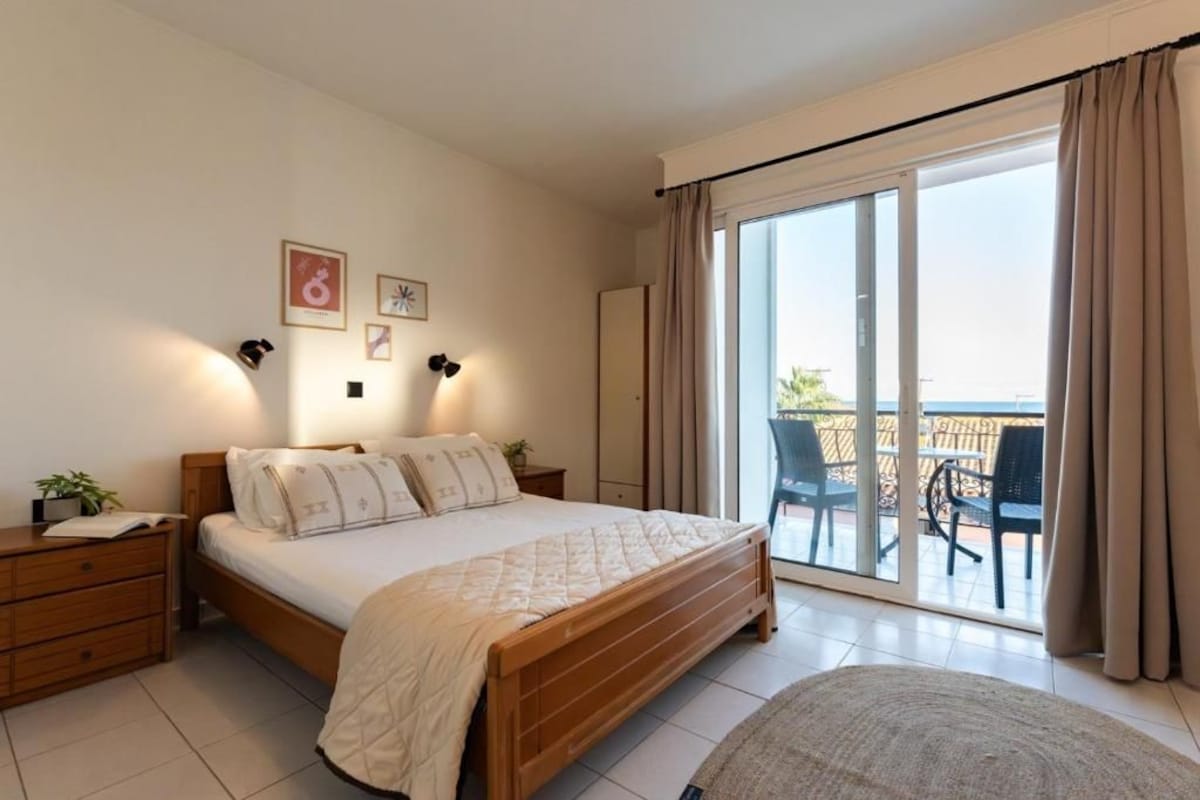 A bedroom with a bed, bedside tables, a nightstand, a window with curtains, and a view of an outdoor balcony.