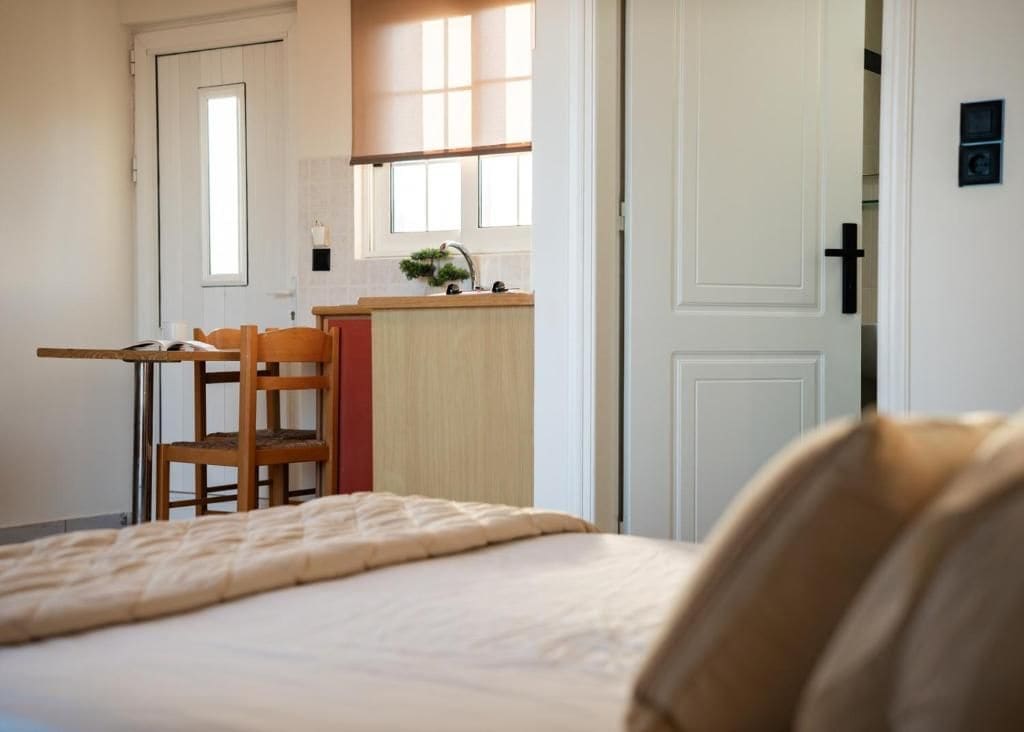 A bedroom with a bed, a wooden chair, a small table, and a kitchenette area with a window.