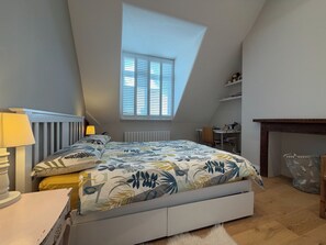 A bedroom with a sloped ceiling, a bed with a floral bedspread, a window with blinds, a bedside table with a lamp, and a small desk with a chair. - Beautiful 1BDR Hideaway in West Hampstead (London)