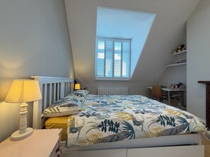 Makuuhuone, jossa on kalteva katto, kattovalaisin, kukkapuvukuvioitu peitto sängyllä, yöpöytälamppu ja ty öpöytä tuolilla.