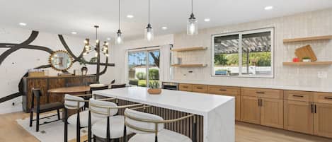 A modern kitchen with a central island, bar stools, pendant lighting, and a large window.