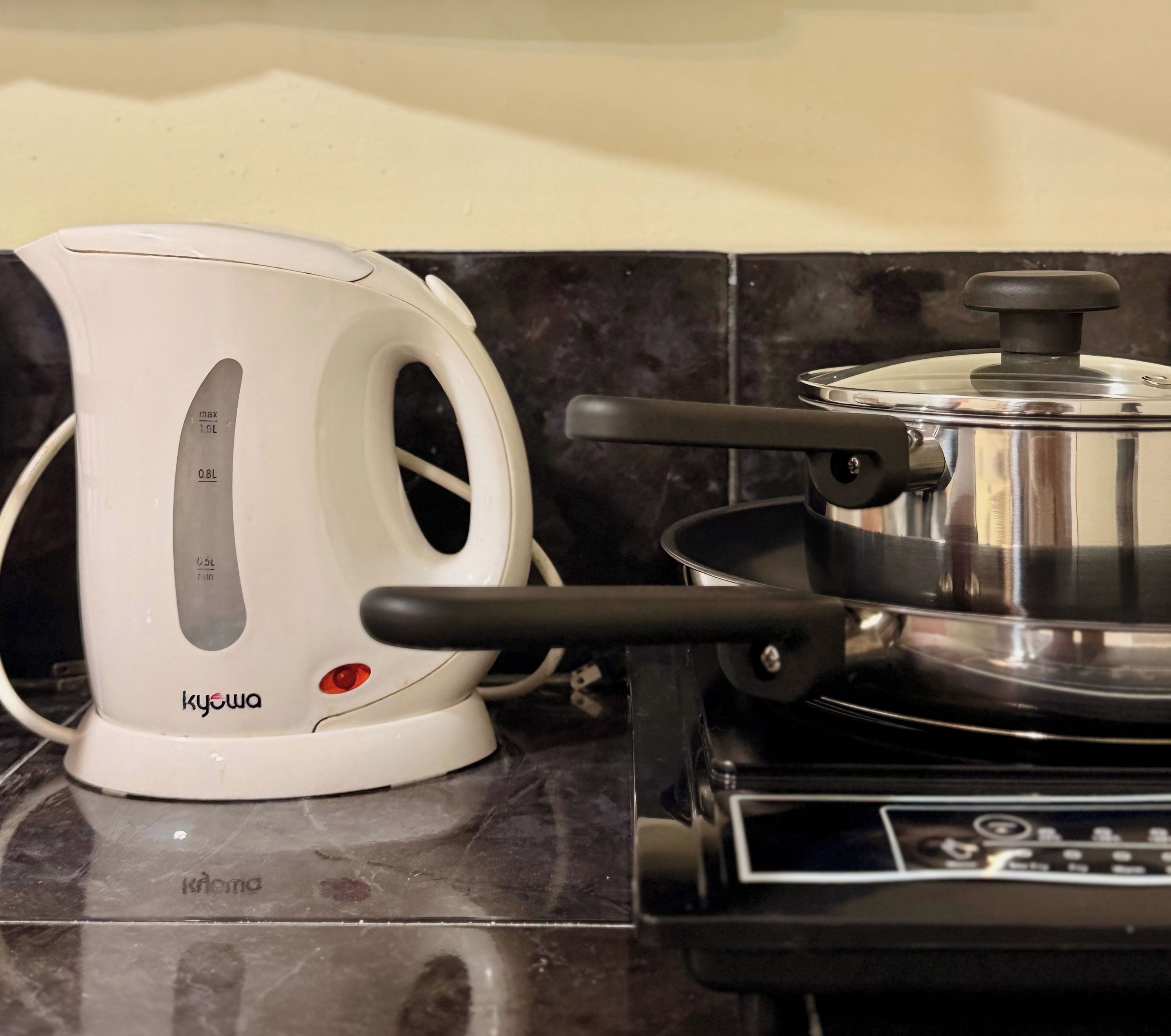 Uma chaleira elétrica branca com luz indicadora vermelha em uma bancada da cozinha, ao lado de um fogão com uma panela.