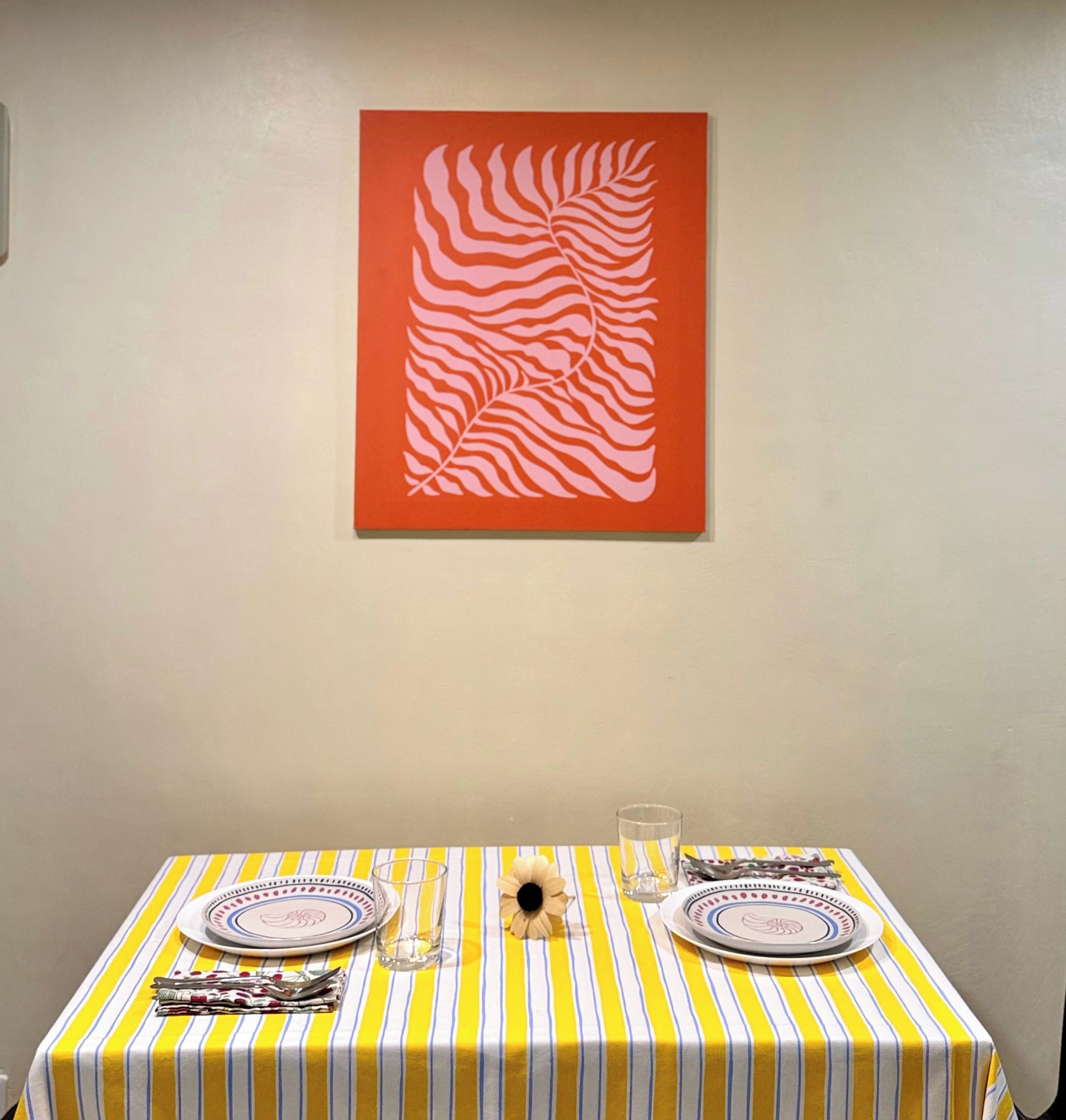 Mesa de jantar com toalha listrada, pratos, talheres e um copo. Uma obra de arte emoldurada está na parede acima.