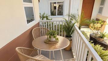 A covered patio with a table and two wicker chairs, surrounded by potted plants and a white picket fence.