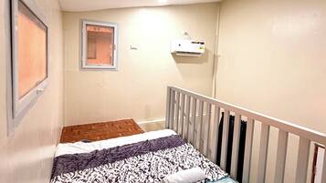 A small, single-bed room with a wooden floor, a white crib, a wall-mounted air conditioner, and a window with a blind.