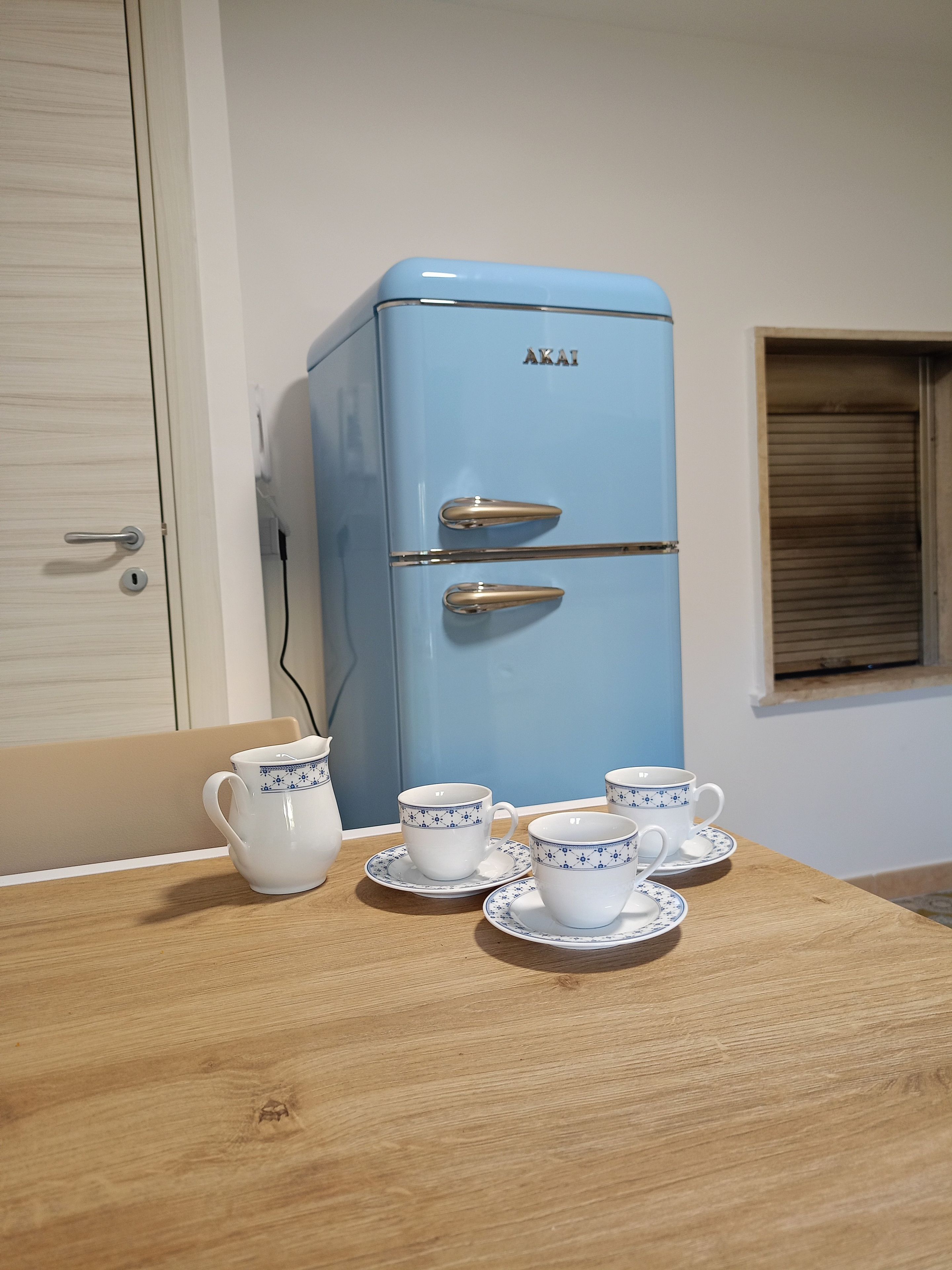 Un réfrigérateur bleu à deux portes, une table en bois avec des tasses et des soucoupes blanches, et une fenêtre avec des stores.