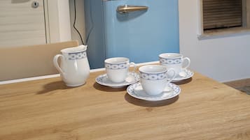 A blue refrigerator with two doors, a wooden table with white cups and saucers, and a window with blinds.