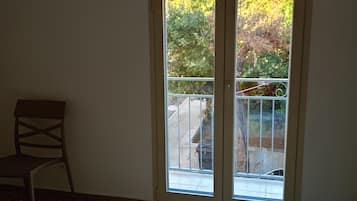 Una habitación con un gran ventanal corredera que da a un balcón, una silla y una cama con cubrecama blanco.