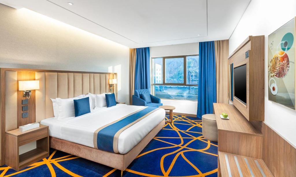 Habitación de hotel con una cama grande, televisor, escritorio y ventana con vistas a un edificio y árboles.