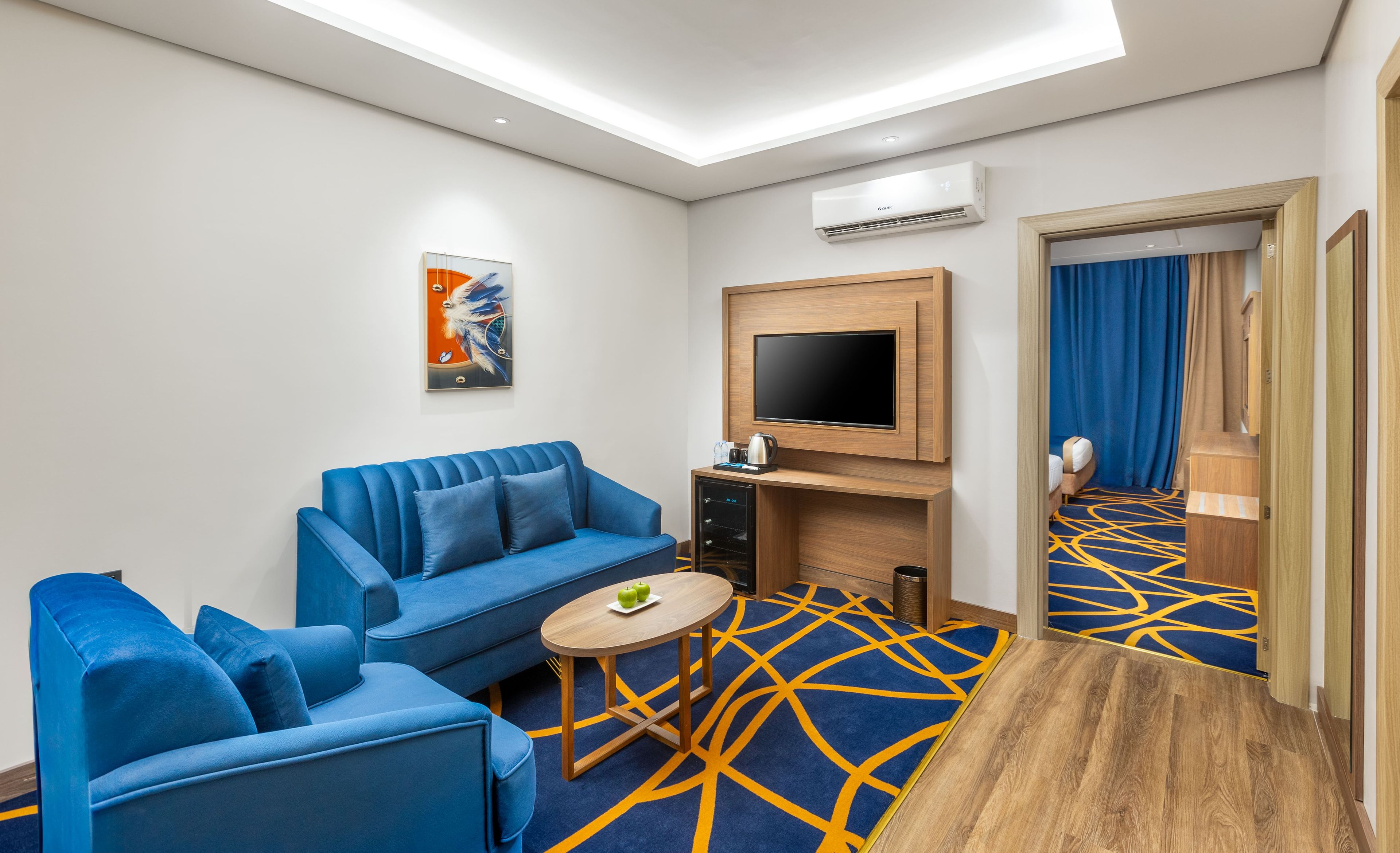 Habitación de hotel moderna con sofá azul, una mesita redonda, televisor de pantalla plana y alfombra estampada.