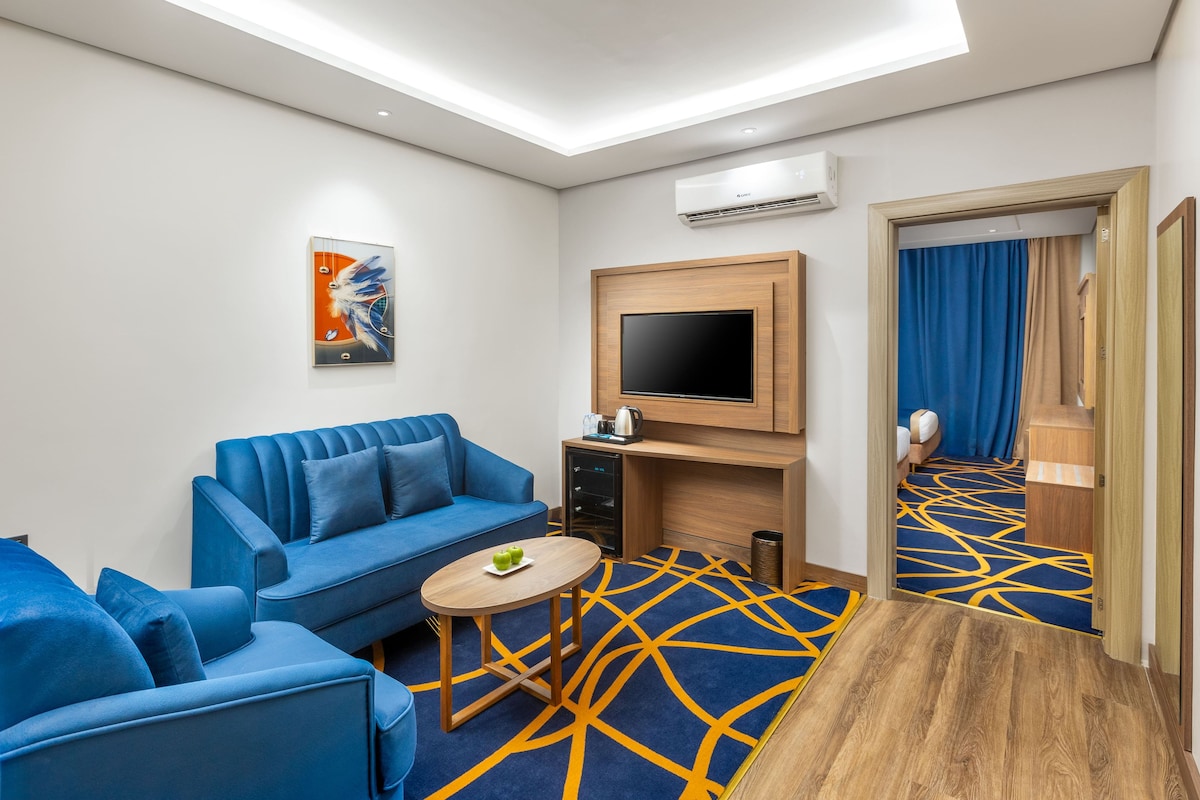 Een moderne hotelkamer met een blauwe zithoek, een klein rond tafeltje, een flatscreen televisie en een kamerbreed tapijt met dessin.
