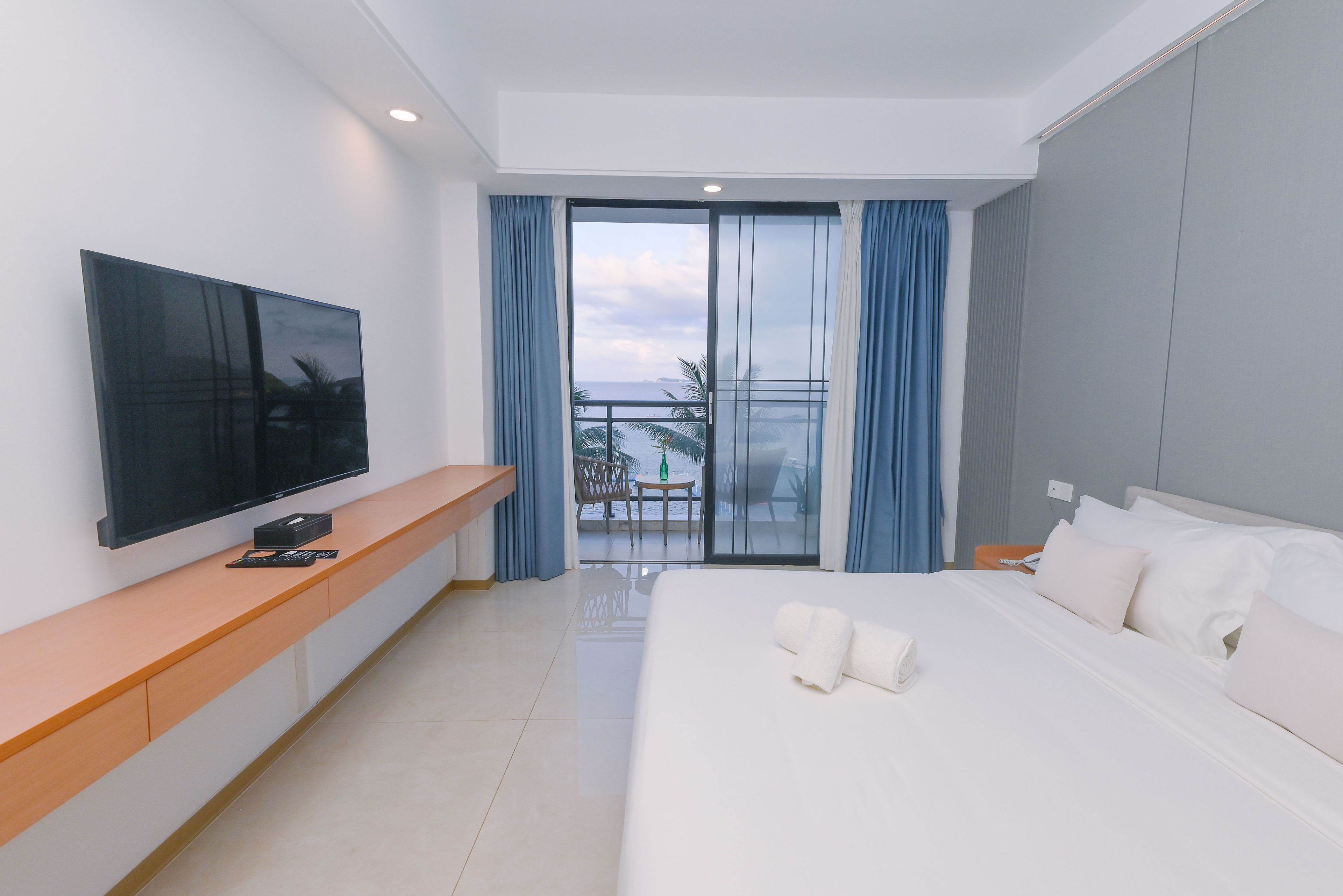 Une chambre d’hôtel avec un lit, un téléviseur posé sur une console en bois, un balcon avec vue et une grande fenêtre avec des rideaux.