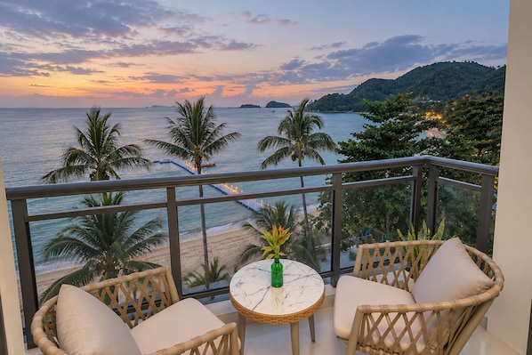 A balcony with wicker furniture, a small round table, and a view of the beach and ocean at sunset.