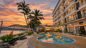 A hotel pool area with lounge chairs, umbrellas, and a view of the sea at sunset.