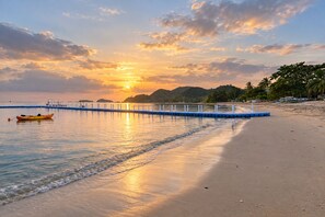 Ein Strand mit einem Steg, der sich in ruhiges Wasser erstreckt, ein Boot am Ufer und ein Sonnenuntergang im Hintergrund.