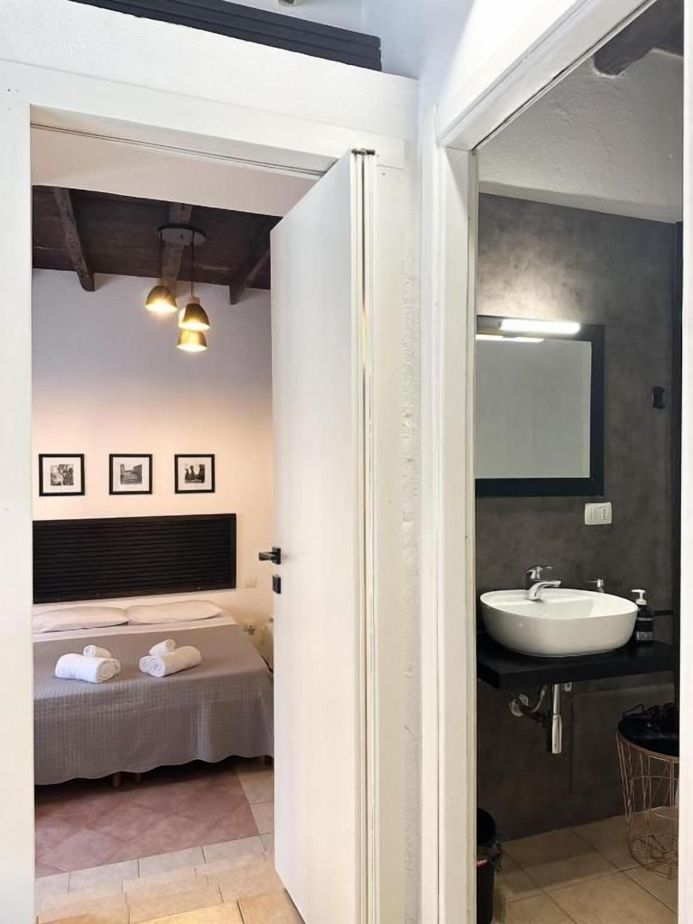 A bedroom with a bed, bedside table, and a nightstand. There is a bathroom with a sink, mirror, and towel rack.