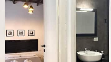 A bedroom with a bed, bedside table, and a nightstand. There is a bathroom with a sink, mirror, and towel rack.