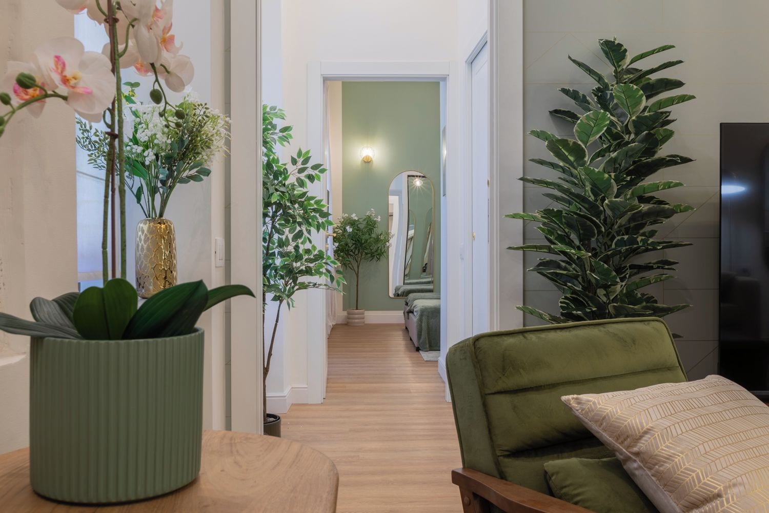 Un pasillo con un sofá verde, una planta en maceta y un espejo que refleja una escalera.